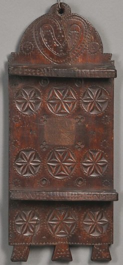 Brown painted carved wooden spoon rack, possibly Bergen County, New Jersey, 1767, with chip carved heart on the arched crest above nine floral medallions and two slotted tiers for eight spoons flanking an indistinctly inscribed label, incised "1767" on the reverse